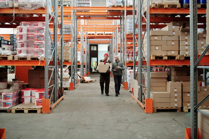 Two workers handle a package in a spacious warehouse surrounded by shelves stocked with boxes and products