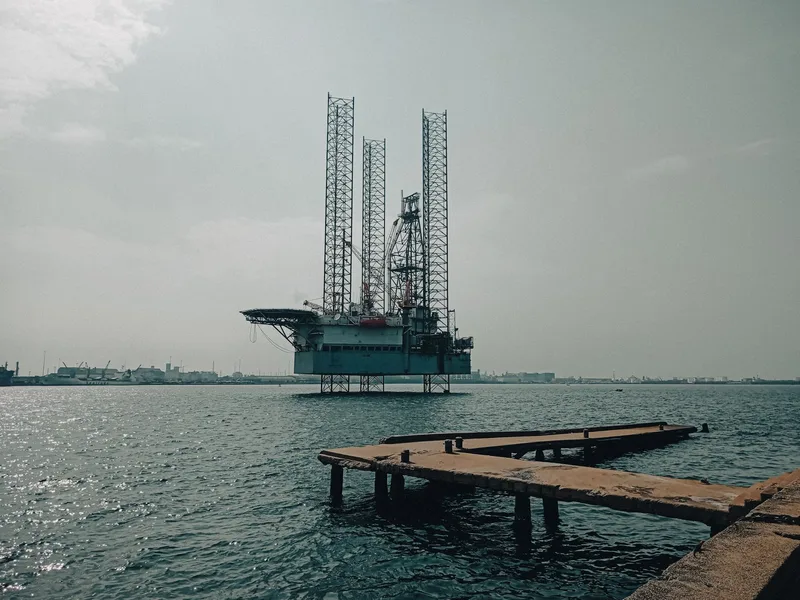 A jack-up offshore drilling rig stands in calm water near a harbour, with a concrete pier in the foreground