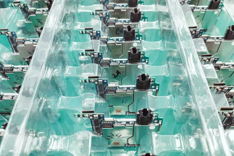 Small circuit boards with headphone jacks sit in clear plastic trays on an electronics production line