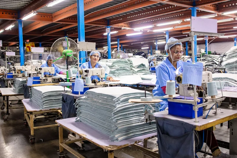 Workers in blue uniforms operate sewing machines beside stacks of cut fabric on a garment factory floor